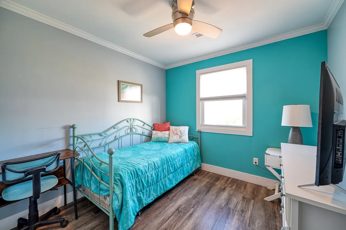 A twin/trundle bedroom featuring a sleek metal bed frame, a bold turquoise accent wall, and a white desk with a chair situated under a ceiling fan. The room is designed for both rest and productivity, with a calm, serene mood.