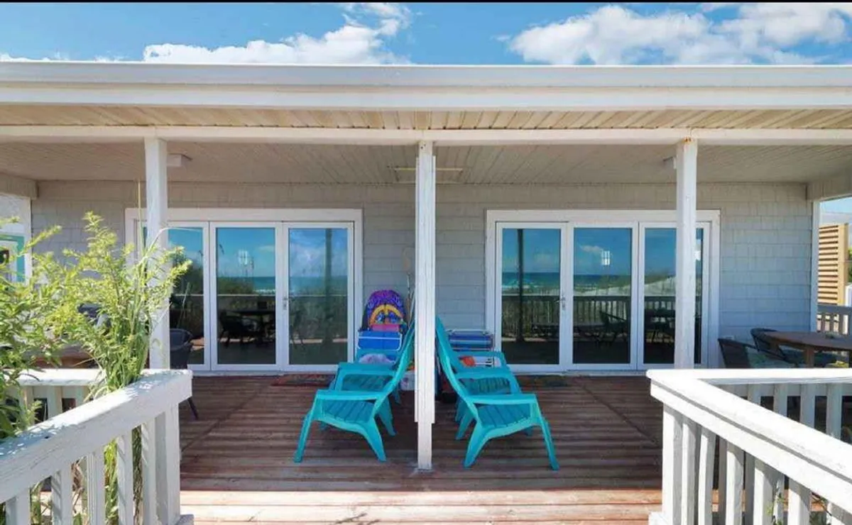 This image presents two turquoise chairs set on a porch overlooking the sea, with sliding glass doors leading back into the home. The scene is bathed in natural light, and the inviting seating area emphasizes a welcoming atmosphere for relaxing and socializing while enjoying the ocean view.