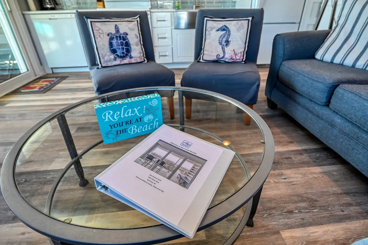 A cozy, beach-themed entry area with comfy chairs, a coffee table, a welcome sign, and a property binder. This interior shot evokes a sense of arrival and hospitality as soon as you enter the home.