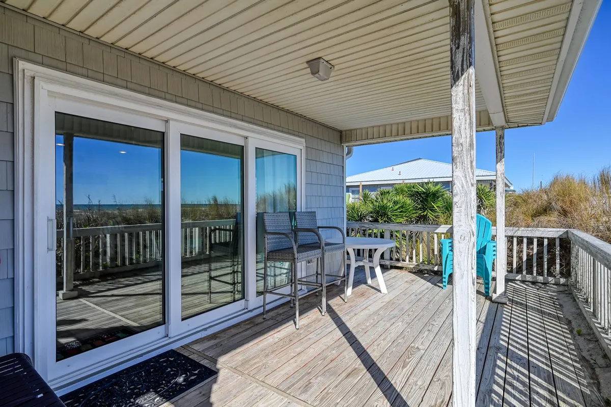 A wide wooden deck with glass doors, inviting outdoor seating, and surrounding vegetation. The deck wraps around the home, offering glimpses of both entry points and relaxing spaces that catch ocean breezes.