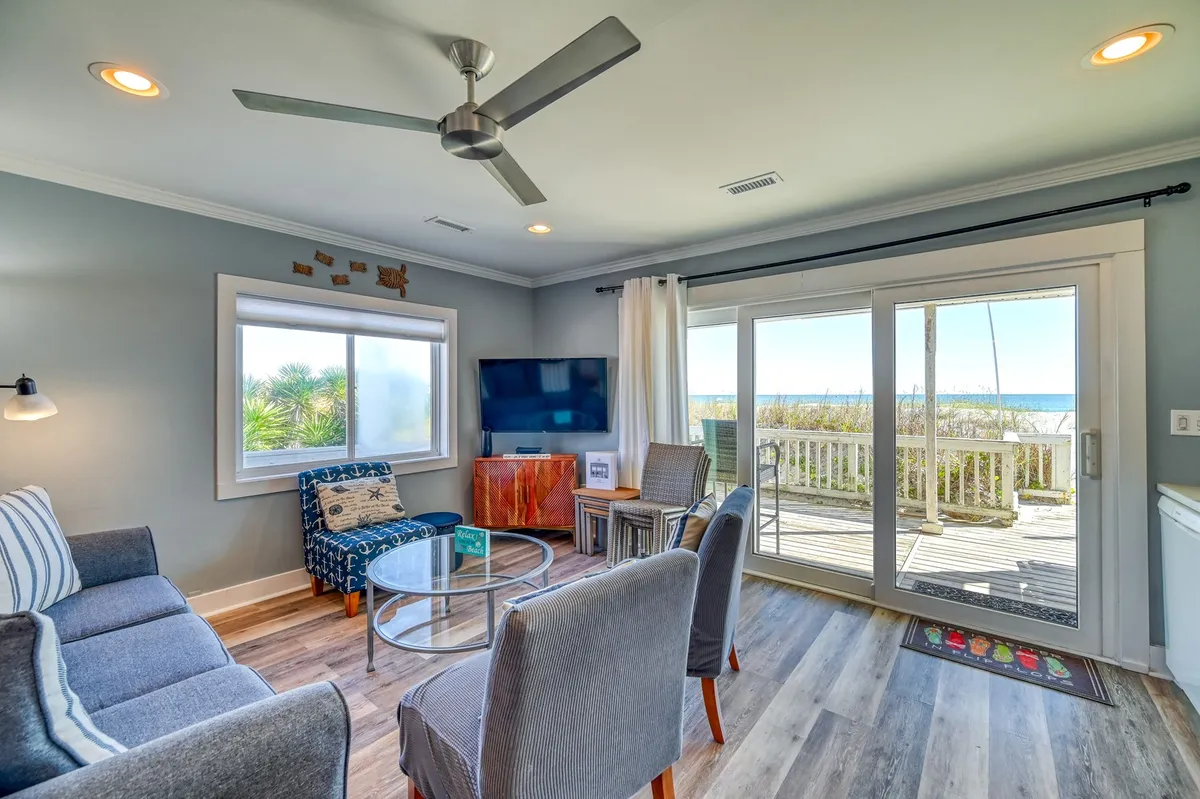 A bright living room with a large window framing stunning ocean views, a comfortable sofa, coffee table, and television. The space is filled with natural sunlight, inviting guests to relax, socialize, or enjoy movie nights while keeping the ocean in sight.
