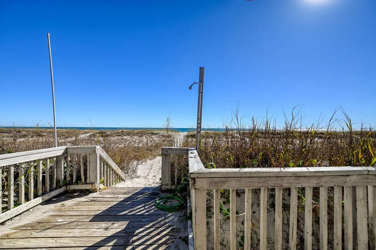 A wooden pathway bordered by beach grass leads directly to the ocean, set against a backdrop of blue sky and glistening water. The scene evokes the feeling of easy, private access from the home to the beach.