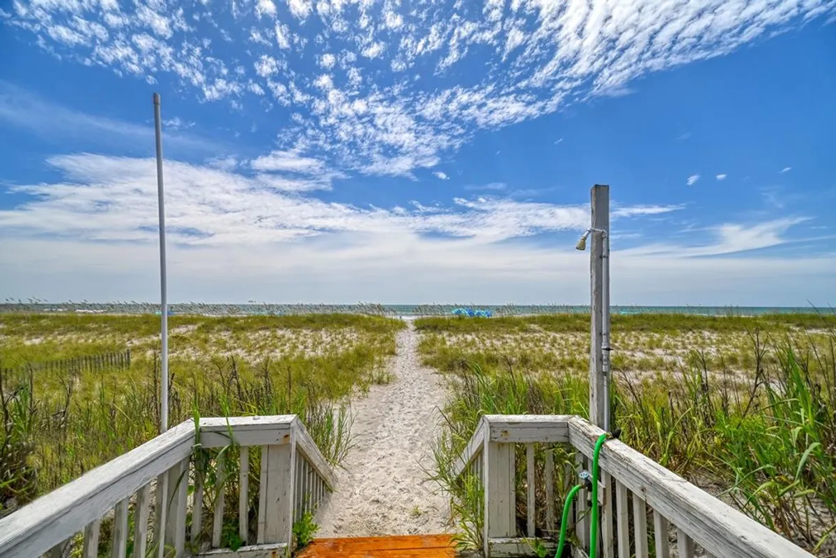 A sandy pathway bordered by tall beach grass leads directly to the ocean, under a vivid blue sky dotted with clouds. The untouched sand dunes and the proximity of the water create an inviting scene of direct, private beach access. There are hints of people enjoying classic beach activities such as walking, sunbathing, and sandcastle building.