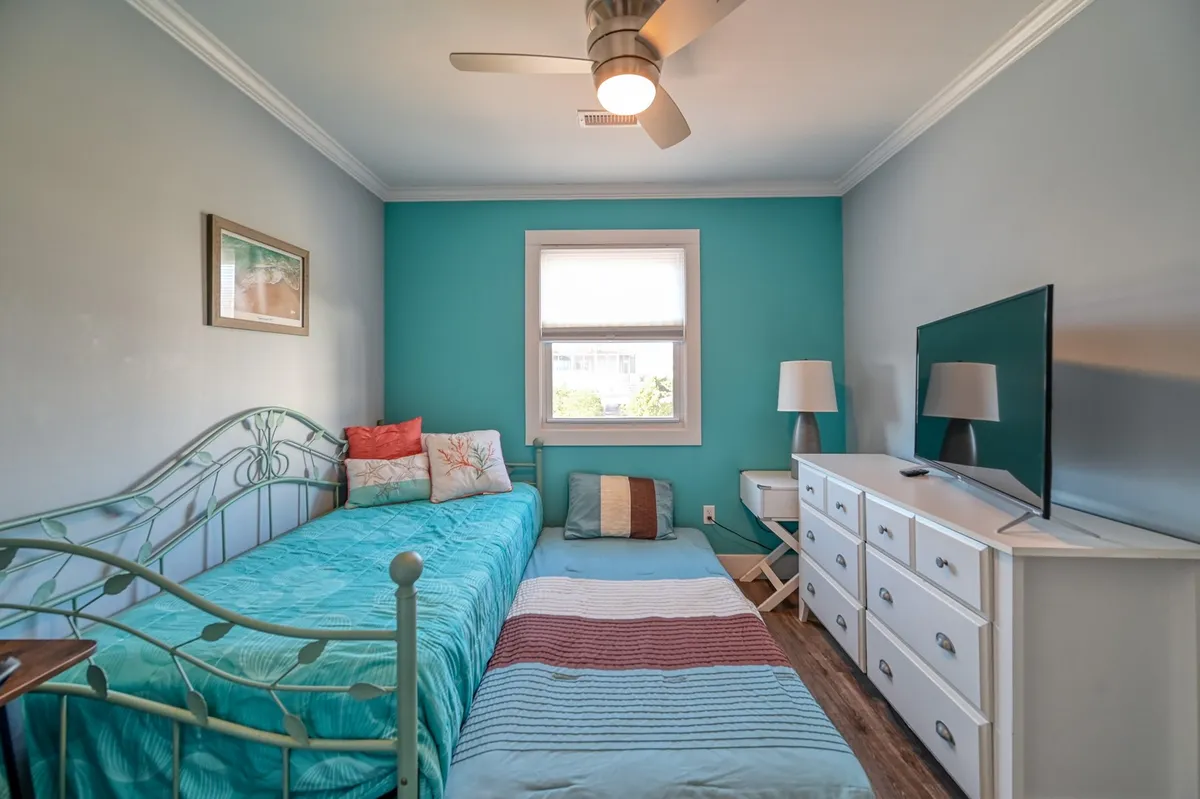 A bright queen bedroom with a comfortable bed decorated with cheerful pillows, a teal accent wall, a white dresser, and a large window letting in abundant natural light. The room exudes a cozy, uplifting atmosphere perfect for relaxation.