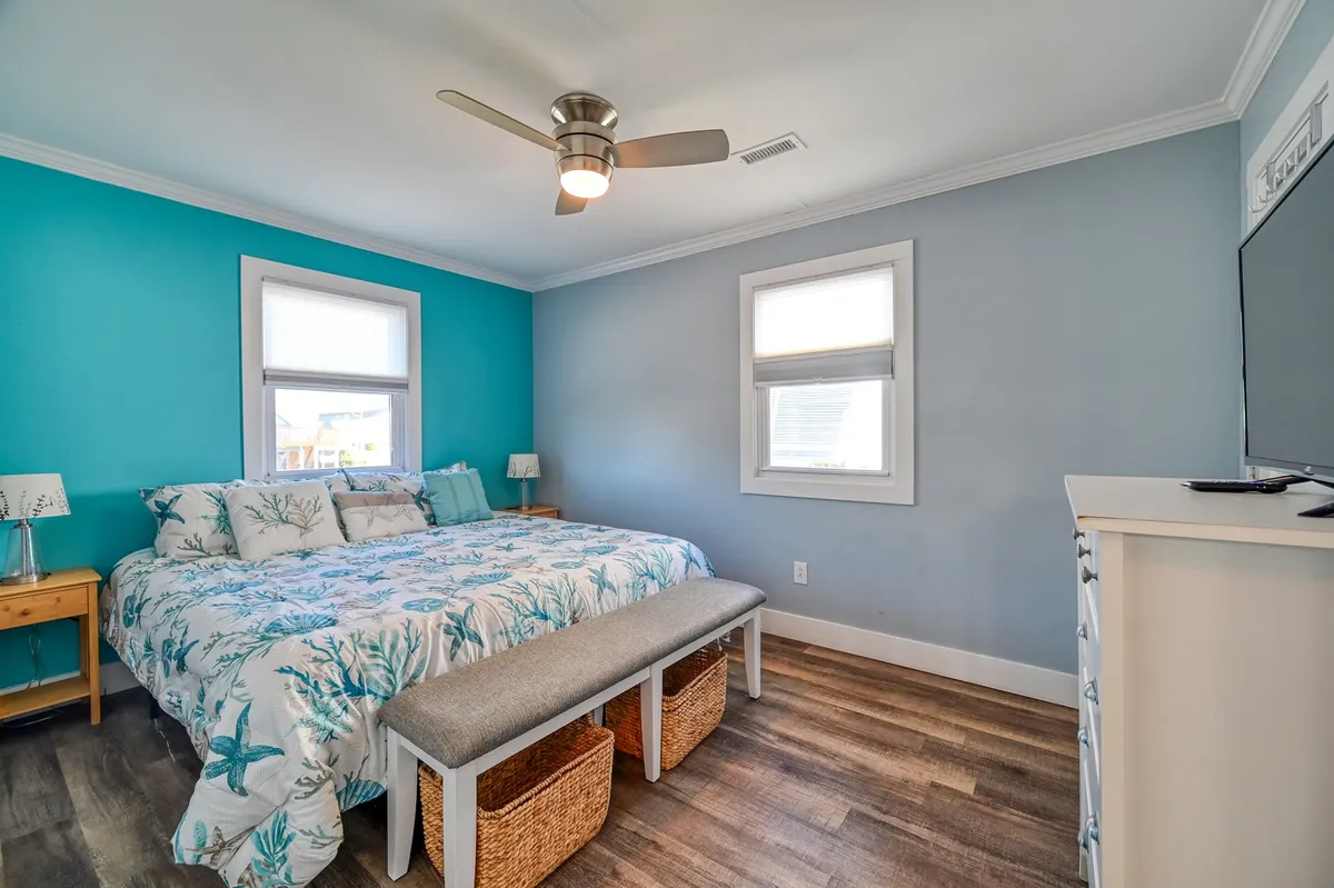 A serene bedroom featuring a plush king bed adorned with floral bedding, a soft blue accent wall, a ceiling fan overhead, and a storage bench with baskets at the foot of the bed. Subtle natural light filters in, highlighting the peaceful, inviting ambiance ideal for restful nights.