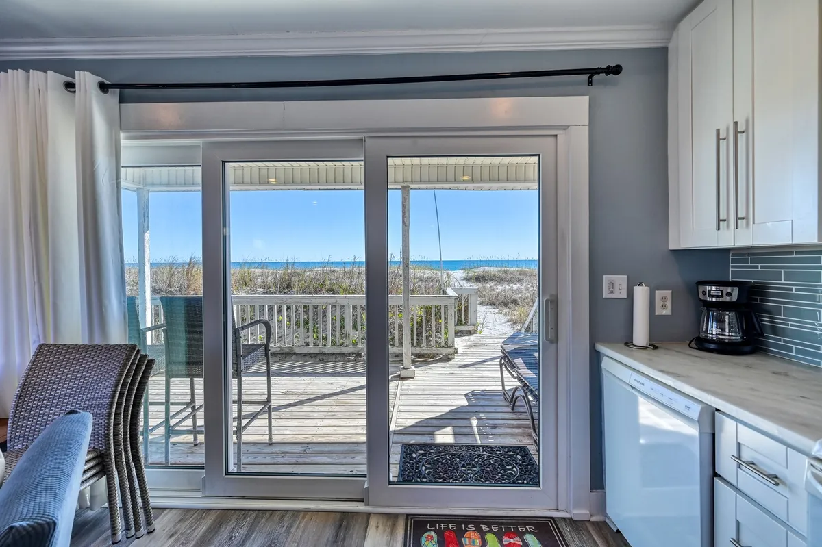 This inviting interior shot centers on a bright, open living space leading to a sliding glass door, which frames a sweeping ocean view. The deck just outside the door promises seamless indoor-outdoor living—perfect for savoring coffee at sunrise or relaxing with a book as gentle waves roll in. Thoughtful details like a coffee maker and a welcoming decorative mat add comfort and warmth.