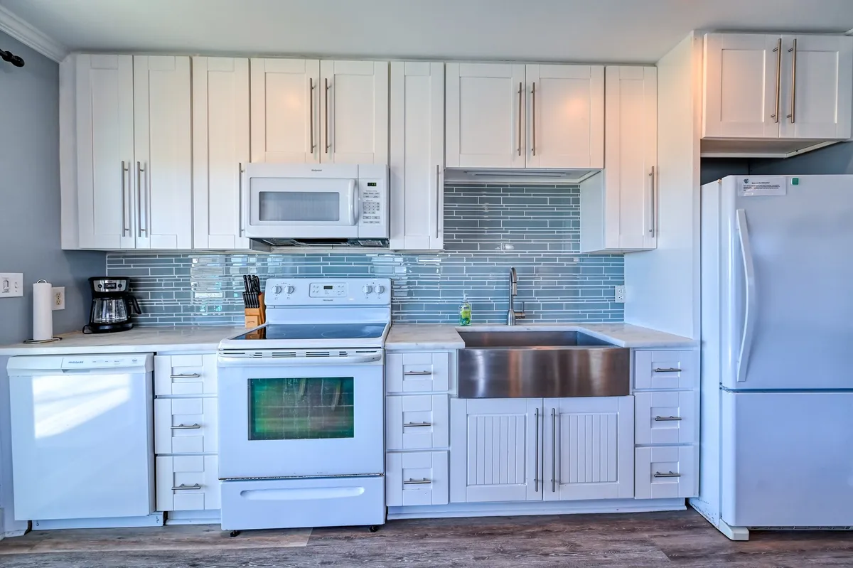 A modern kitchen space equipped with a stainless steel farmhouse sink, blue tile backsplash, white cabinetry, and countertop appliances. The design is both functional and stylish, showcasing custom tilework and updated features ideal for cooking and entertaining.