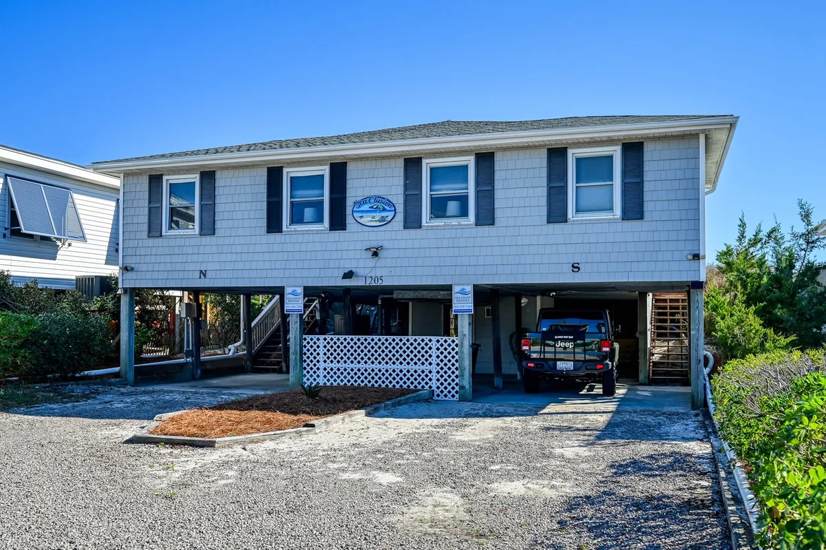 This exterior shot showcases the house structure, visible driveway, and surrounding greenery. The view hints at the elevated positioning of the property and offers a glimpse of the welcoming porch area, tying into the 'seaside welcome' theme.