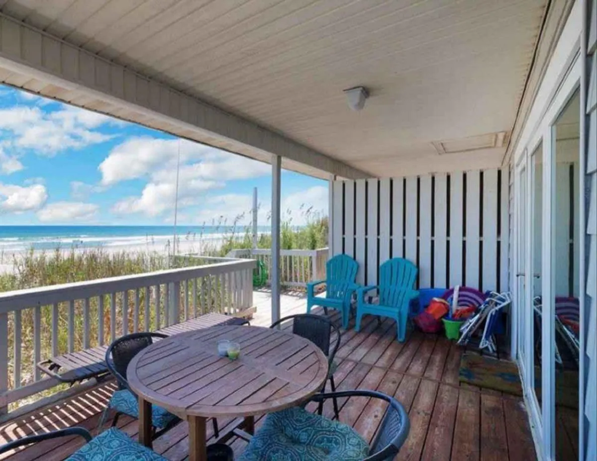 A welcoming dining area featuring a wooden table surrounded by blue Adirondack chairs, overlooking the ocean through expansive windows. The setting encourages gatherings over meals with a breathtaking backdrop and offers a seamless blend of comfort, style, and coastal atmosphere.