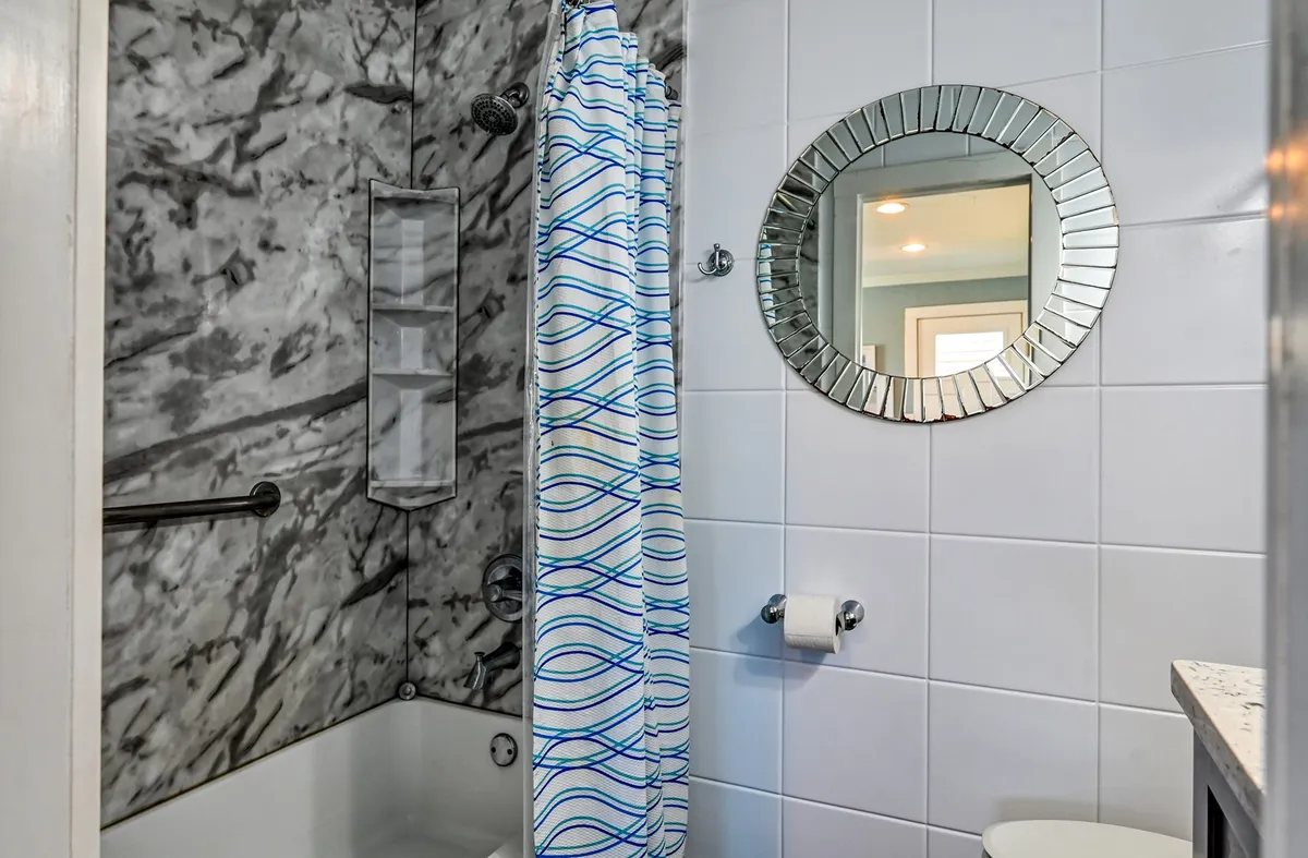 A modern, inviting bathroom with a striking marble wall, a circular mirror above the sink, and a shower curtained with blue waves pattern. The design is fresh and stylish, creating a spa-like atmosphere for guests to refresh.