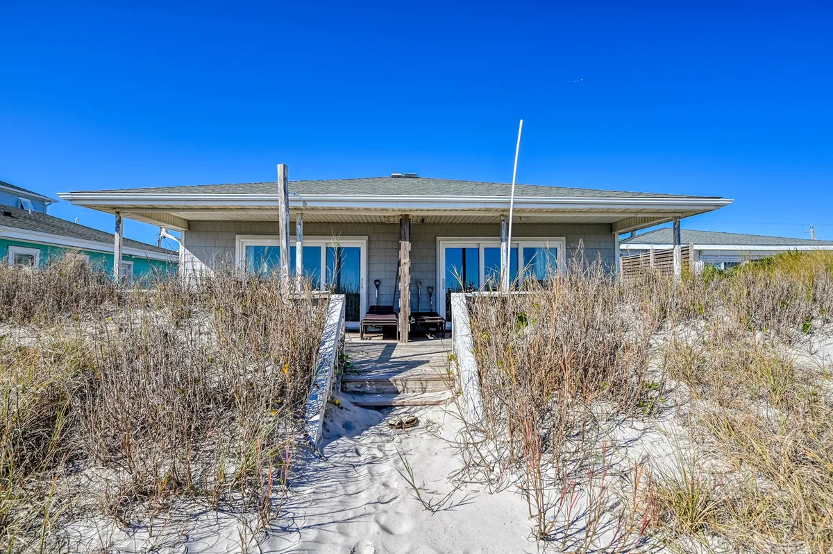 The exterior of a charming beach house is shown, with a sand pathway leading through native vegetation toward the porch. The house sits just behind the dunes, emphasizing its proximity to the ocean. The setting is calm, with soft morning or evening light, and suggests a welcoming home base for beach adventures.